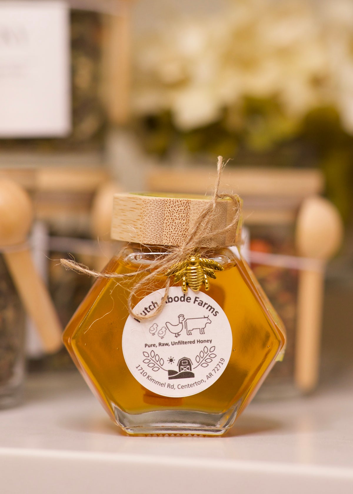 A hexagonal glass jar filled with golden honey, featuring a light wood lid tied with twine and a round white label that reads &