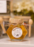 A hexagonal glass jar filled with golden honey, featuring a light wood lid tied with twine and a round white label that reads &